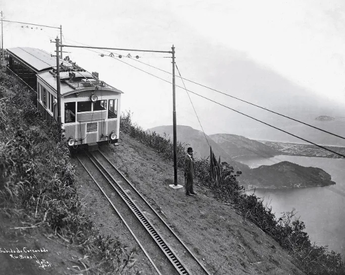 Antigo trem elétrico subindo os trilhos do Corcovado com a Baía de Guanabara ao fundo, foto histórica