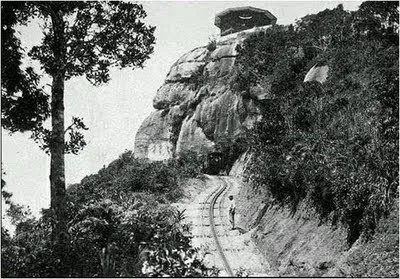 Trem entrando em um túnel sob um grande rochedo, com os trilhos serpenteando pela mata, foto histórica