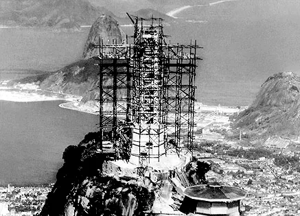 Estátua do Cristo Redentor em construção, coberta por andaimes, com o Pão de Açúcar ao fundo, foto histórica