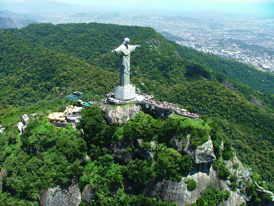 Vista aérea do Cristo Redentor no alto do Corcovado, cercado pela floresta da Tijuca, com a cidade do Rio de Janeiro ao fundo